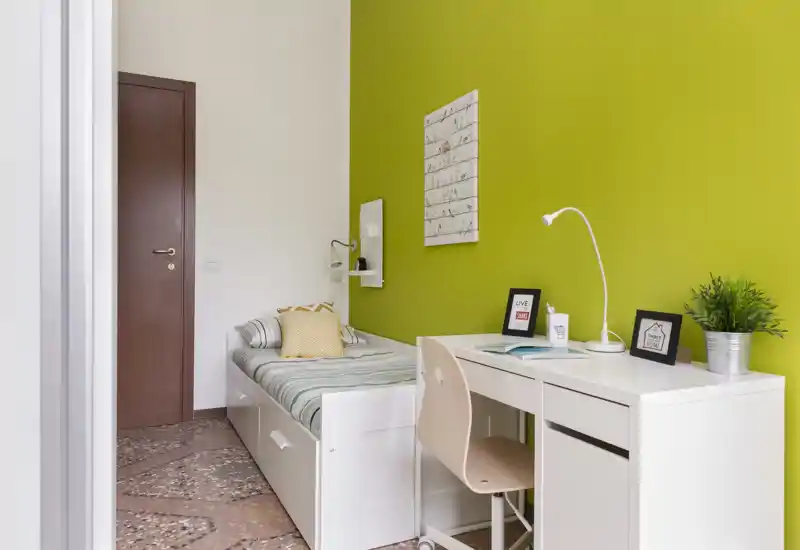 Habitación individual compacta y luminosa con cama blanca con cajones, escritorio y pared de acento verde llamativo — ideal para estudiante o persona sola.