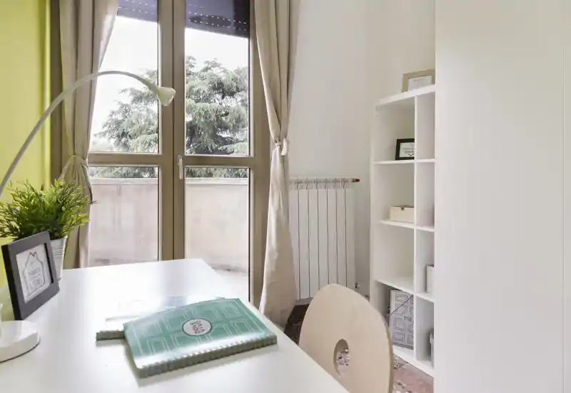 Bright, styled living/working corner with a desk by a large window, neutral curtains and organized shelving — ideal for a comfortable workspace with natural light.