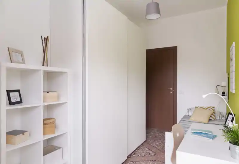 Dormitorio compacto con cama individual, escritorio, estanterías blancas y una pared verde de acento. Espacio luminoso y ordenado, ideal para un estudiante o una sola persona.