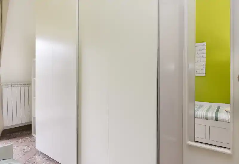 Dormitorio luminoso centrado en un gran armario blanco; un espejo estrecho refleja parcialmente una cama con ropa de cama a rayas. La foto destaca el almacenamiento y un vistazo del área de descanso.