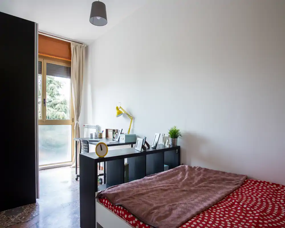 Camera da letto luminosa e semplicemente arredata con letto matrimoniale in primo piano, un mobile basso che funge da divisorio e una scrivania vicino alla finestra che riceve luce naturale.