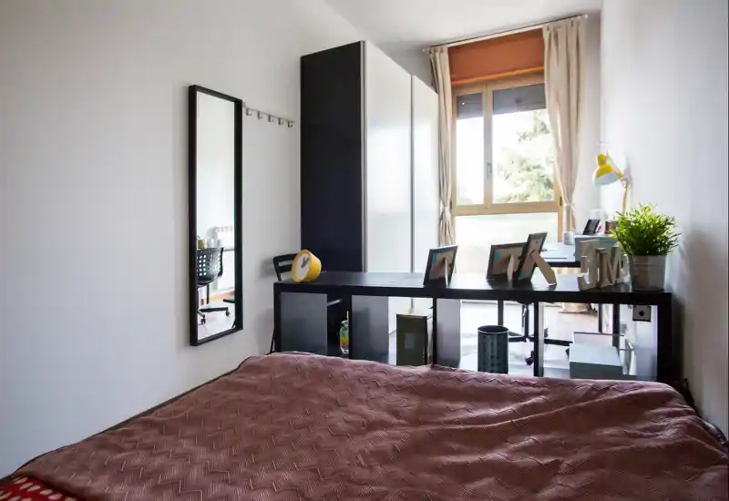 Chambre lumineuse et soignée avec un grand lit au premier plan, une étagère derrière le lit, un bureau près de la fenêtre et une agréable lumière naturelle.