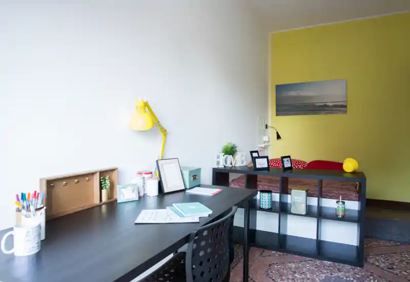 Coin chambre lumineux et moderne avec bureau : bureau et chaise noirs, lampe de bureau, étagère en cubes et lit simple contre un mur jaune. L'espace est décoré avec de petits objets, adapté pour étudiants ou locations courtes durées.