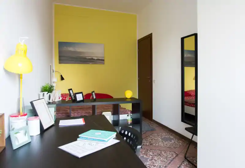 Coin chambre moderne et lumineux avec un bureau au premier plan, une étagère basse séparant l'espace nuit et un lit bien fait à la literie rouge devant un mur d'accent jaune.