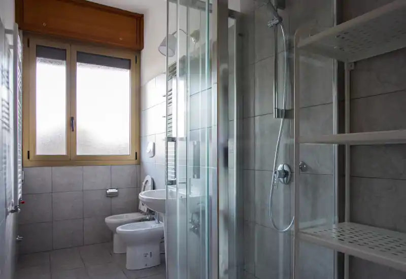 Bright, modern bathroom with tiled walls, a glass shower enclosure, bidet, toilet and sink under a frosted window.