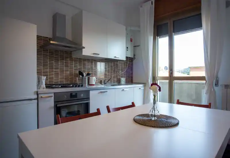 Modern fitted kitchen with white cabinets, stainless-steel appliances and a bright dining table by a large window – ideal for a sunny breakfast spot.