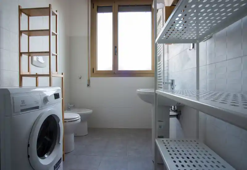 Bright, clean bathroom with a washing machine, toilet, bidet and sink beneath a large window; practical shelving offers storage.