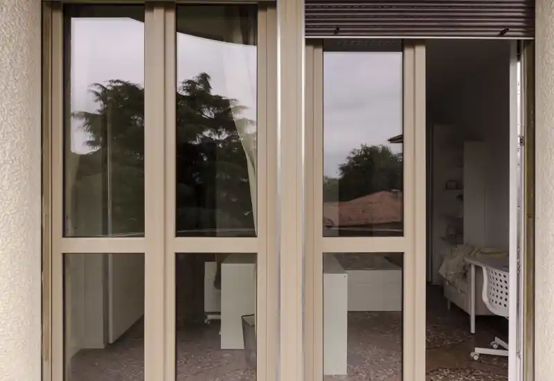 Vista attraverso porte scorrevoli in vetro su una camera da letto piccola con letto parzialmente visibile, sedia da scrivania e scaffalatura. La luce naturale entra dalle porte, creando un’atmosfera calma e neutra.