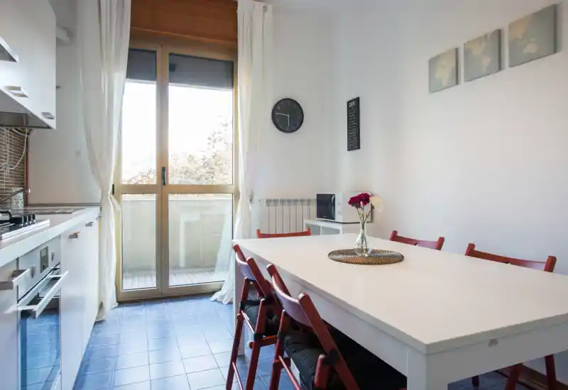 Bright modern kitchen with a large white dining table, red folding chairs and a balcony door that fills the room with natural light. Clean styling, tiled floor and practical appliances make it ideal for short-term rentals.