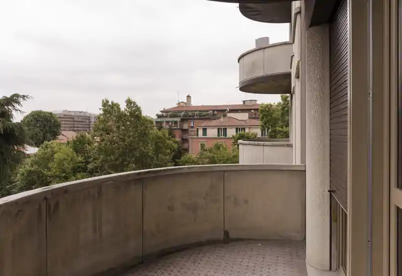 Sunny-looking curved balcony with a tiled floor and a view over trees and nearby buildings — a private outdoor space suitable for morning coffee.