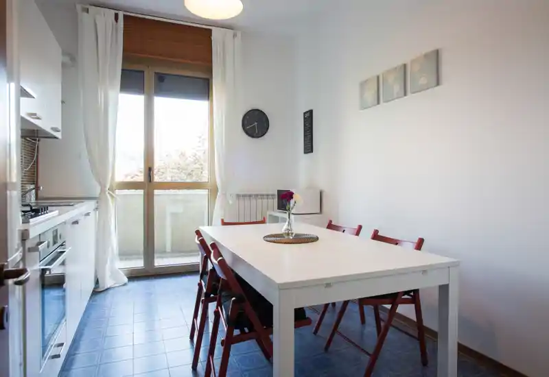 Bright, modern kitchen with an integrated dining area: white cabinets, a large window letting in natural light, and a simple white dining table with red folding chairs and a vase of flowers.