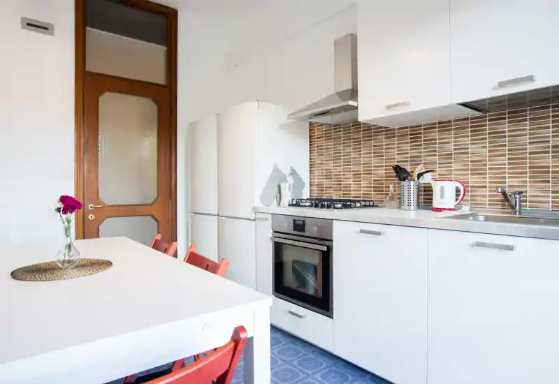 Bright modern kitchen with white cabinets, a built-in oven, gas hob, fridge and a small dining table with red chairs — well-lit and neatly styled for rental listings.