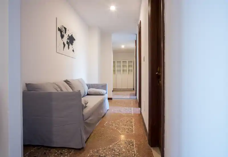 Compact living area with a grey upholstered sofa, soft cushions and minimal wall art; clean, well-lit hallway view leads toward a cabineted room, ideal for cozy apartment listings.