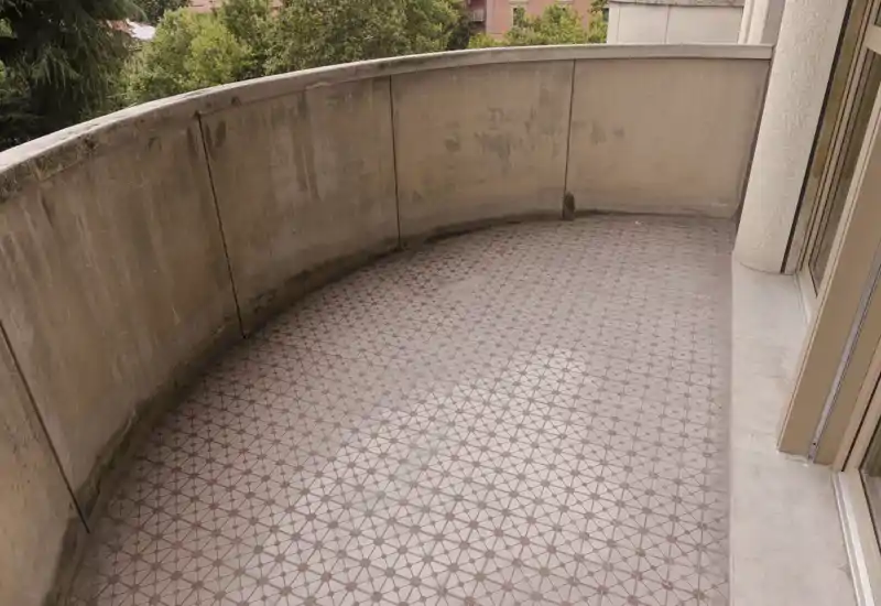 Small curved balcony with patterned tiled floor and low concrete balustrade; view overlooks trees and neighboring buildings. Ready for seating or plants with direct access from the interior.