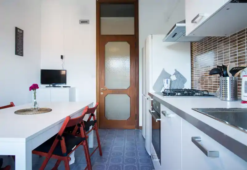 Bright, compact kitchen with an integrated dining area: white cabinets, gas hob, tiled backsplash and a simple table with red folding chairs.