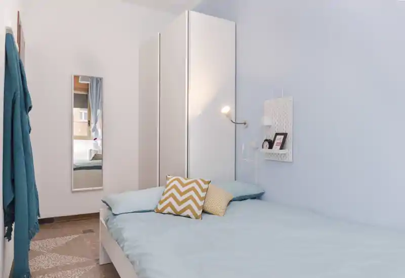 Chambre lumineuse et soignée avec un lit double en linge bleu pâle, coussins décoratifs et lampe murale. Une armoire étroite et un miroir plein pied agrandissent l'espace.