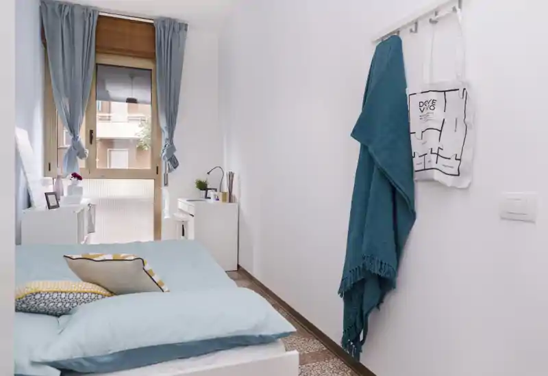 Chambre lumineuse et soignée avec linge de lit bleu pâle, coussins décoratifs et un petit bureau près de la fenêtre — chaleureuse et bien mise en valeur pour une annonce.