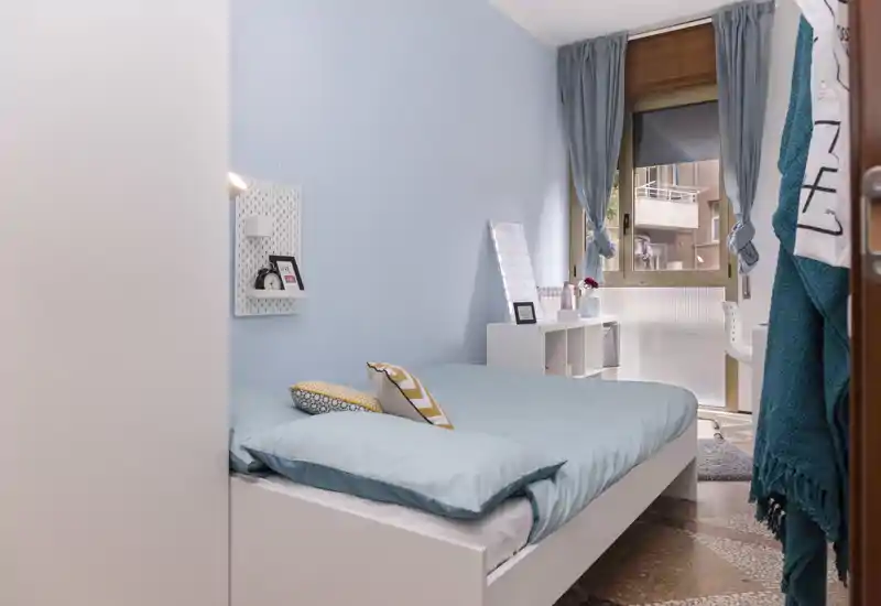 Chambre lumineuse et soignée avec un lit double en linge bleu clair, coussins décoratifs et une fenêtre apportant de la lumière naturelle. Un petit bureau et des étagères près de la fenêtre forment un coin pratique et chaleureux.