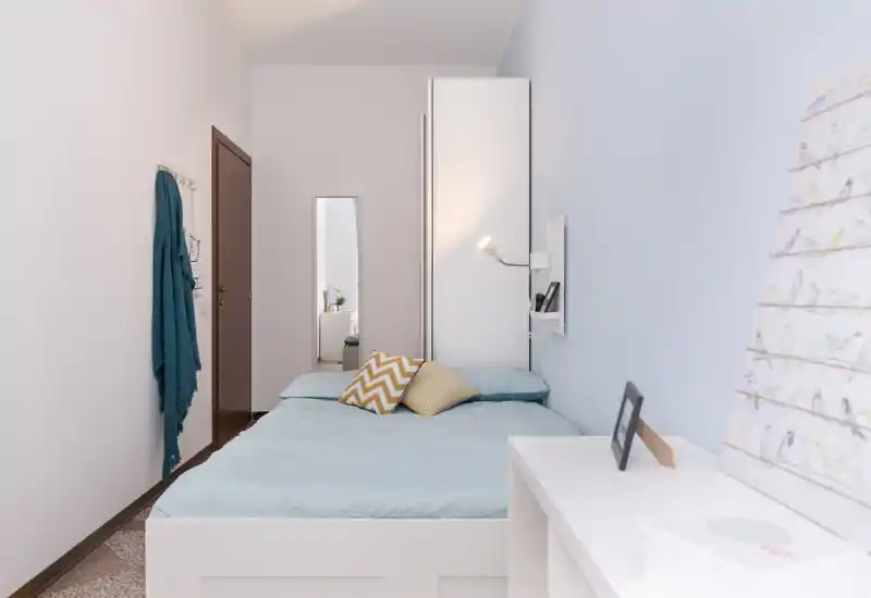 Chambre compacte et lumineuse avec un lit bien fait, coussins à motifs et murs bleu pâle offrant une ambiance sereine et minimaliste.