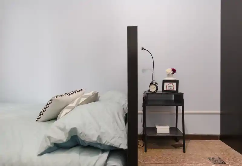 Coin chambre lumineux et moderne avec lit visible habillé de linge pastel et coussins décoratifs, à côté d'une petite table de chevet noire avec lampe et réveil.