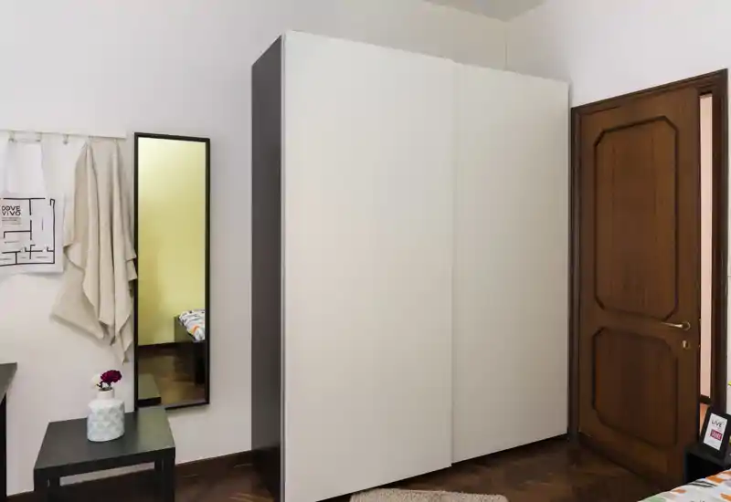 Compact bedroom corner with a large white wardrobe, full-length mirror and a wooden door; the bed appears only as a partial reflection in the mirror. Clean, modern styling with parquet floors and soft accents.