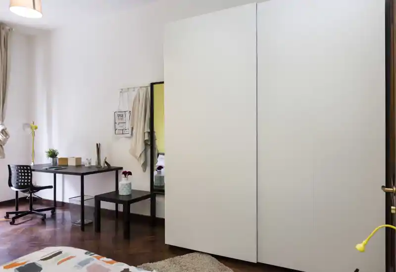 Rincón de dormitorio luminoso y ordenado con borde de la cama visible, gran armario blanco y un pequeño puesto de trabajo junto a la ventana—ideal para una persona o estudiante.