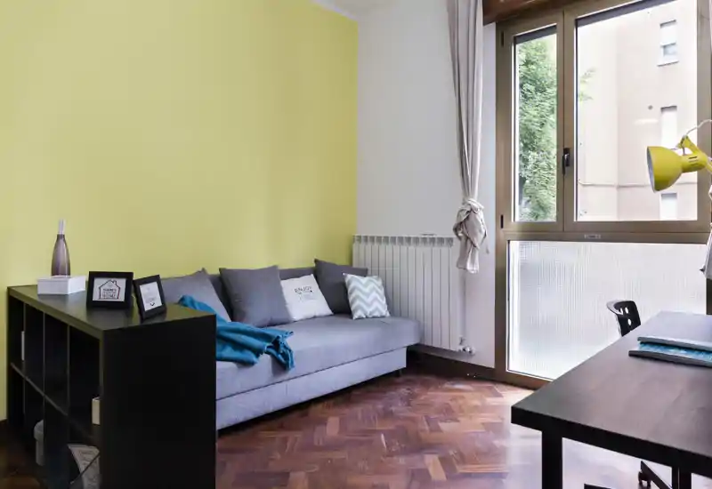 Bright, tidy living area with a grey sofa, decorative cushions and a yellow accent wall. Large window provides natural light and a workspace is visible at the right.