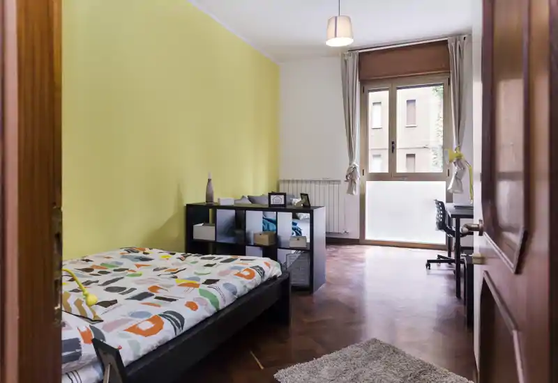 Dormitorio luminoso y ordenado con cama individual, ropa de cama colorida, estantería de cubos, escritorio junto a la ventana y pared amarilla.