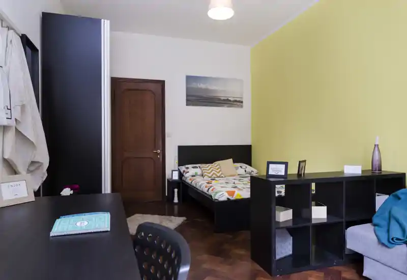 Dormitorio luminoso y bien decorado con cama doble, ropa de cama estampada y detalles acogedores como alfombra y cojines. Incluye pared amarilla, estantería y escritorio para trabajar o almacenamiento.