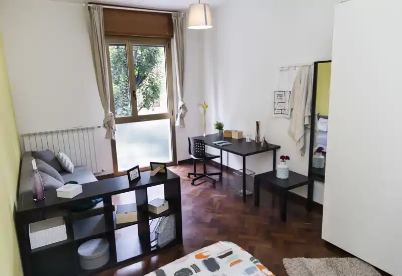 Dormitorio luminoso y ordenado con colcha estampada en primer plano, un pequeño rincón de trabajo junto a la ventana y estanterías que delimitan la zona de descanso.