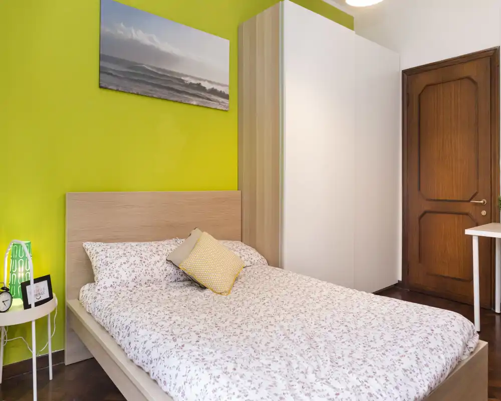 Camera da letto luminosa e accogliente con letto matrimoniale, biancheria a fantasia, comodino e ampia armadiatura bianca. La parete verde e il quadro marino donano personalità e un tocco moderno.