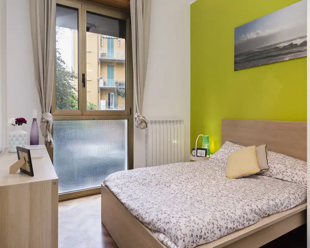 Camera da letto luminosa e accogliente con letto matrimoniale, copriletto fantasia e parete verde a contrasto; ampia finestra con luce naturale.