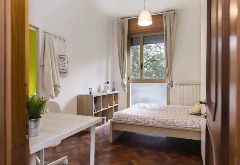 Dormitorio acogedor y luminoso con cama de madera y ropa de cama estampada, ventana con cortinas que aporta luz natural y mobiliario minimalista.