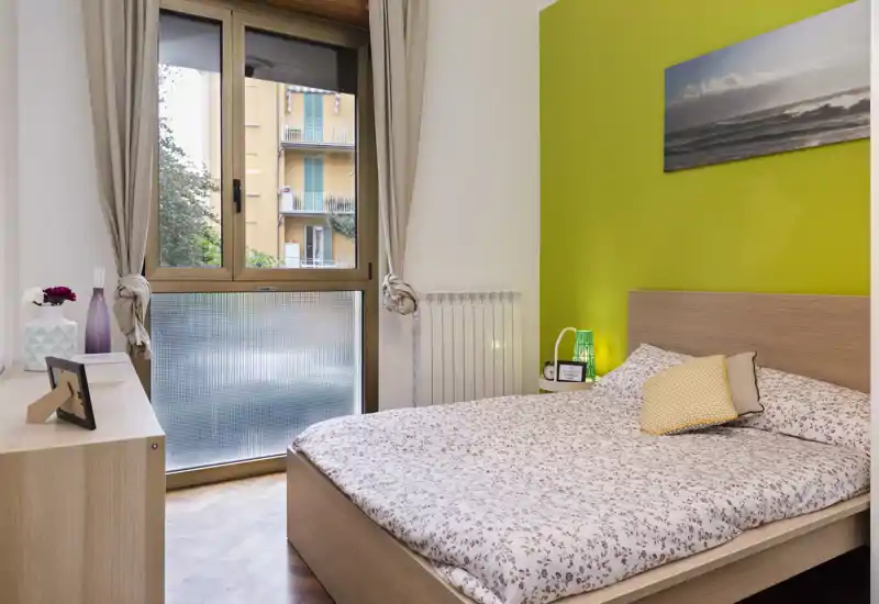 Dormitorio luminoso y acogedor con cama doble, edredón estampado y pared verde; gran ventana que aporta luz natural.