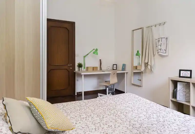 Dormitorio luminoso y acogedor con colcha floral, cojines decorativos y una mesa de trabajo sencilla — ideal para descansar y trabajar ligeramente.
