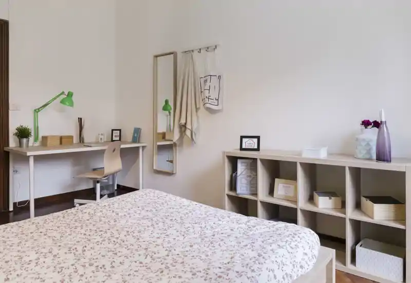 Dormitorio luminoso y acogedor con edredón floral, escritorio con lámpara verde, espejo y estantería organizada — ideal para estudiantes o profesionales que buscan una habitación privada ordenada.