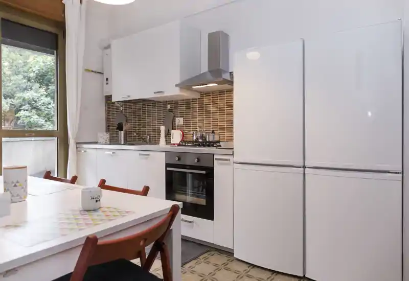 Bright modern kitchen with white cabinets, built-in oven, gas hob and a double-door fridge. A small dining table with colorful placemats and chairs sits by the window, creating a practical and welcoming cooking area.