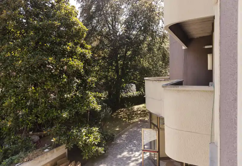 Sunny balcony view overlooking a leafy garden with mature trees and a paved pathway, offering a pleasant outdoor space.