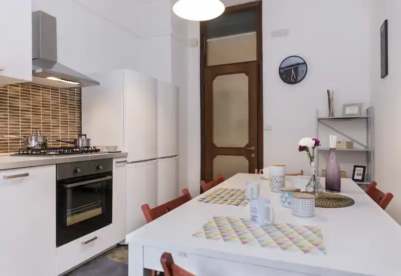 Bright, modern kitchen with a built-in oven, gas hob and full-height fridge beside a dining table set for four. Clean white cabinetry, decorative placemats and fresh flowers create a welcoming, functional space ideal for everyday meals.