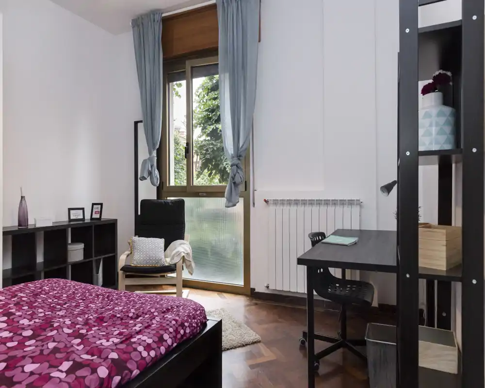 Camera da letto luminosa e ordinata con letto matrimoniale a fantasia, poltrona vicino alla finestra e scrivania funzionale — ideale per riposo e studio, con luce naturale.