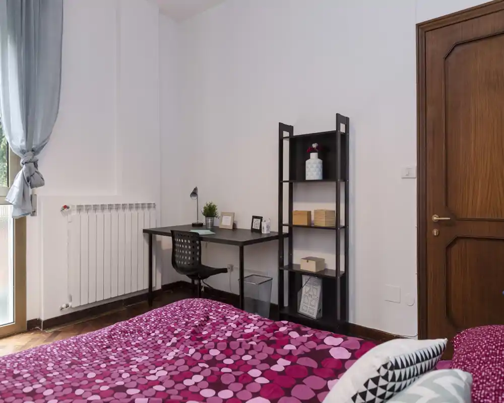Camera da letto luminosa e ben arredata con copriletto fantasia fucsia, scrivania e scaffale ideali per uno studente o viaggiatore. La luce naturale dalla finestra rende l’ambiente arioso e accogliente.