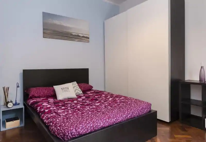 Camera da letto luminosa e ordinata con letto matrimoniale con biancheria fucsia a fantasia, grande armadio e comodino curato — ideale per un soggiorno accogliente.