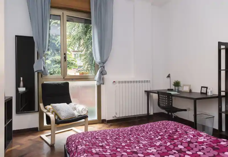 Camera da letto luminosa e ben arredata con copriletto a fantasia, una comoda poltrona vicino alla finestra e una scrivania per lavorare — perfetta per ospiti o studenti.