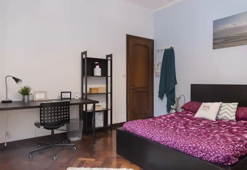 Bright, well-styled bedroom with a double bed dressed in a patterned magenta duvet, decorative pillows and a dark headboard. A workspace with a desk and shelving occupies the left side, and wooden parquet flooring adds warmth.