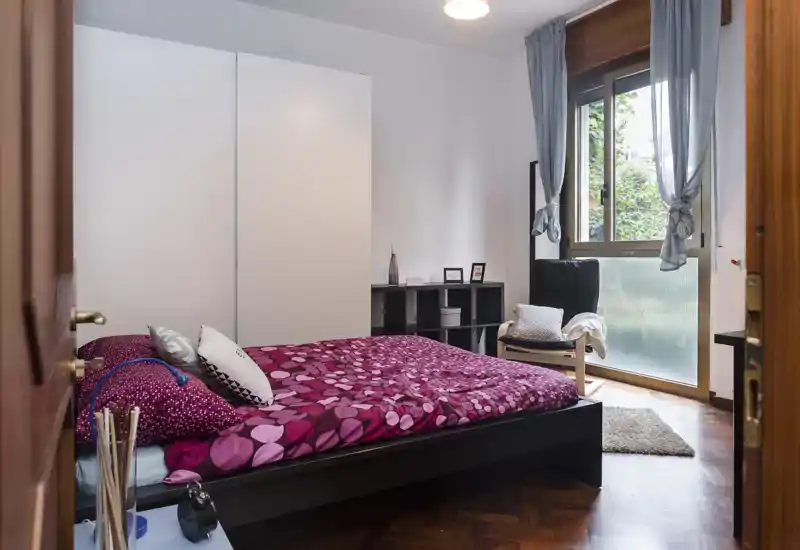 Camera da letto luminosa e ben arredata con piumone viola a fantasia, armadio, poltrona vicino alla finestra e pavimento in legno lucido — perfetta per annunci che evidenziano comfort e luce naturale.