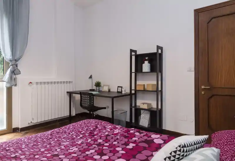 Camera da letto luminosa e ben arredata con copriletto fantasia fucsia, scrivania e scaffale ideali per uno studente o viaggiatore. La luce naturale dalla finestra rende l’ambiente arioso e accogliente.