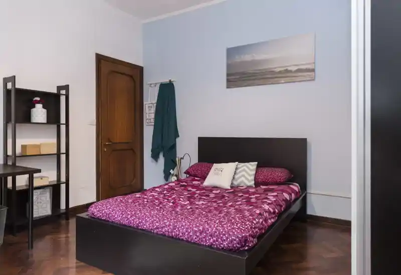 Camera da letto luminosa e ben arredata con letto matrimoniale, biancheria rosa a fantasia, cuscini decorativi e mobili minimalisti per un soggiorno accogliente.