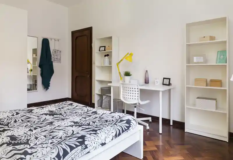 Camera da letto luminosa e ordinata con letto matrimoniale decorato, scrivania bianca e scaffali — ideale per riposo e lavoro da remoto.