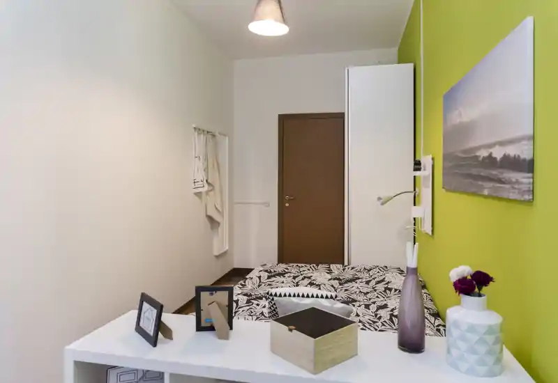 Piccola camera da letto ben illuminata con letto matrimoniale dal copriletto a motivi, parete accentuata verde, armadio e accessori decorativi su una mensola bianca.