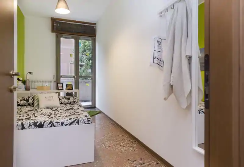 Camera da letto compatta e ben arredata con letto singolo a fantasia, comodino e luce naturale dalla finestra/terrazzino — luminosa e accogliente per un ospite.